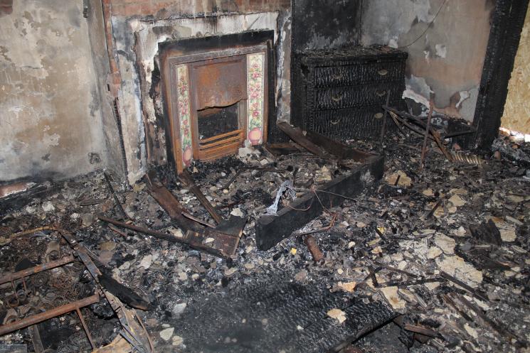 A room inside of the house, with a fireplace and chest of drawers, all completely burned, and debris all over the floor.