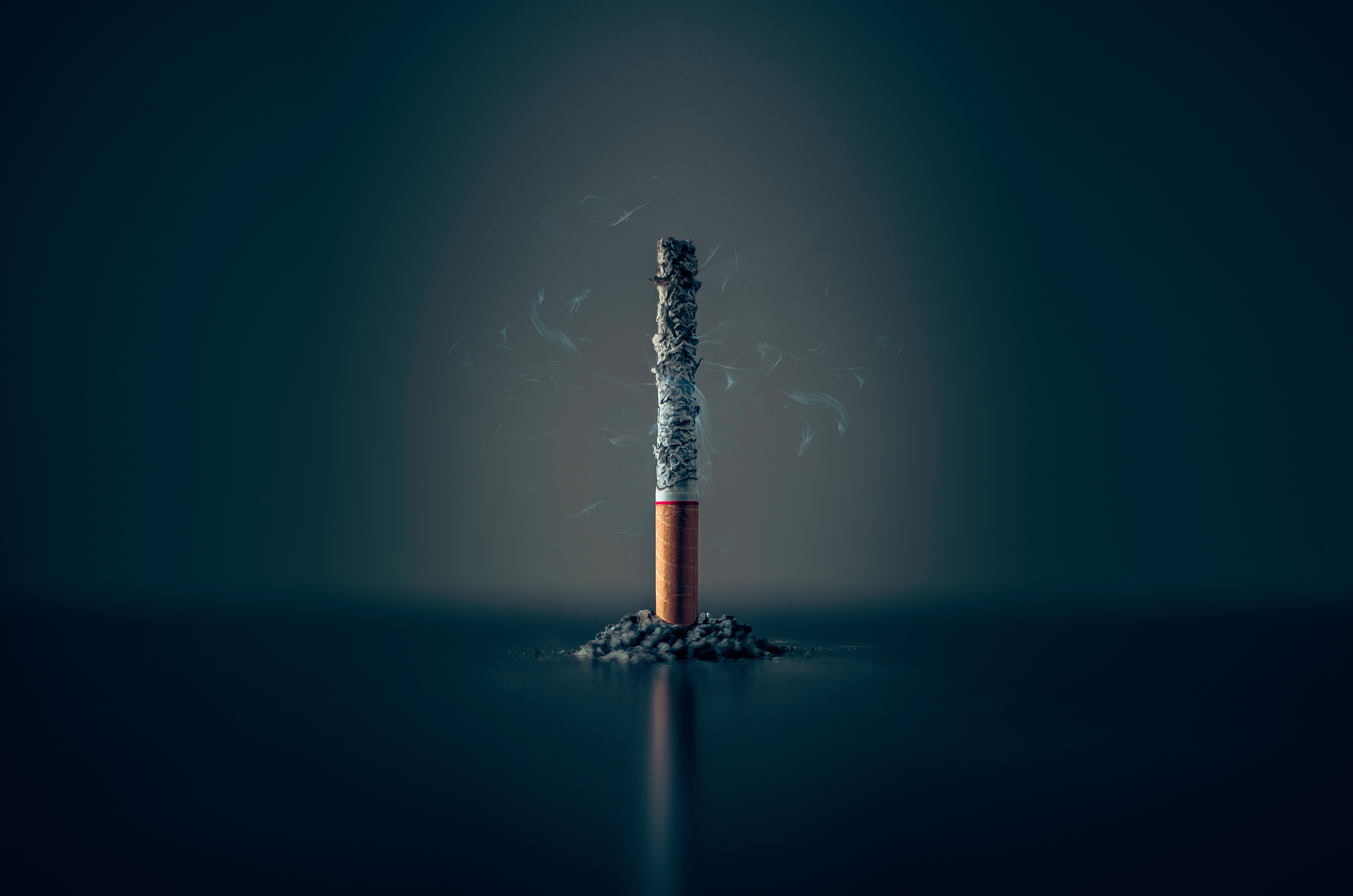 A single lit cigarette with a grey, dark background and a pile of ash below it.