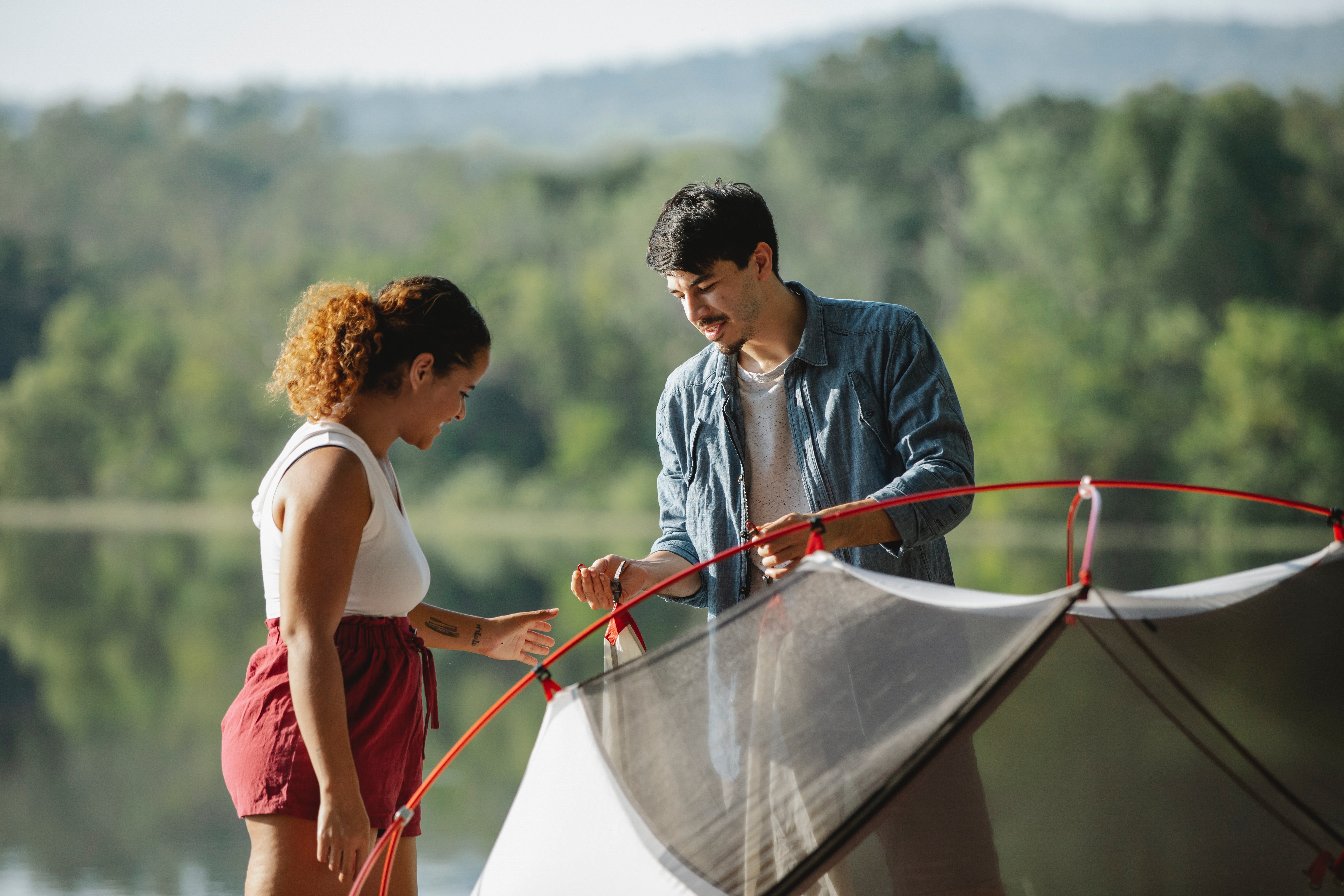 A woman and man setting up a tent together, with a lake and trees in the background.