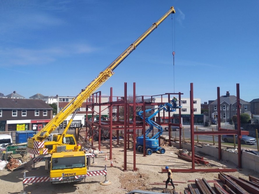 The building site in Plymstock. There's a yellow crane lorry and red frame work. You can also see a blue vehicle which has extended up to the top of the frame work so a builder can work up there.