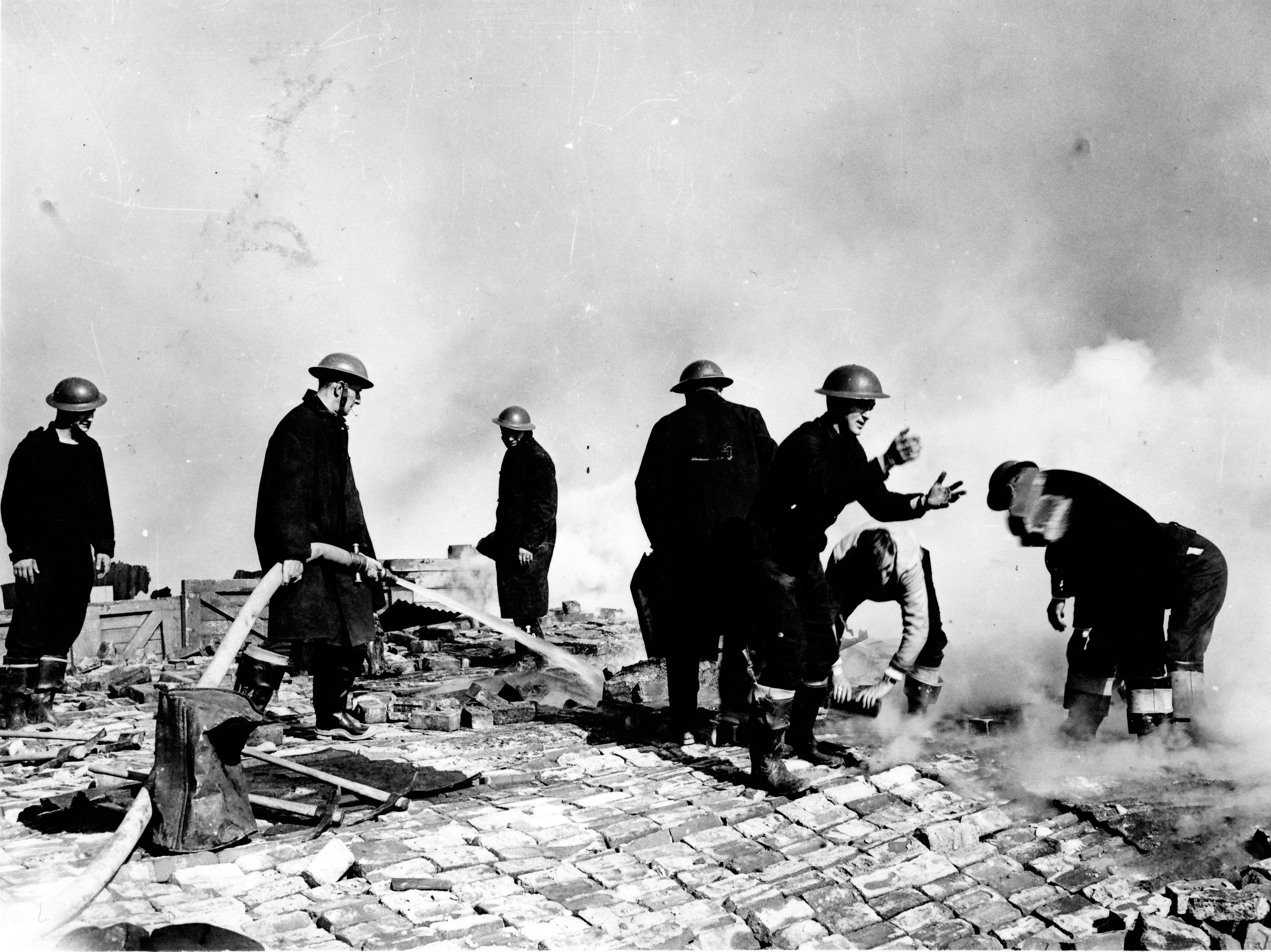 What looks to be eight firefighters in the streets, which are ablaze. They are in full uniform (jumpsuits and helmets). One is holding a hose, one is reaching to catch something, two are stood looking around and others are crouched down handling bricks.