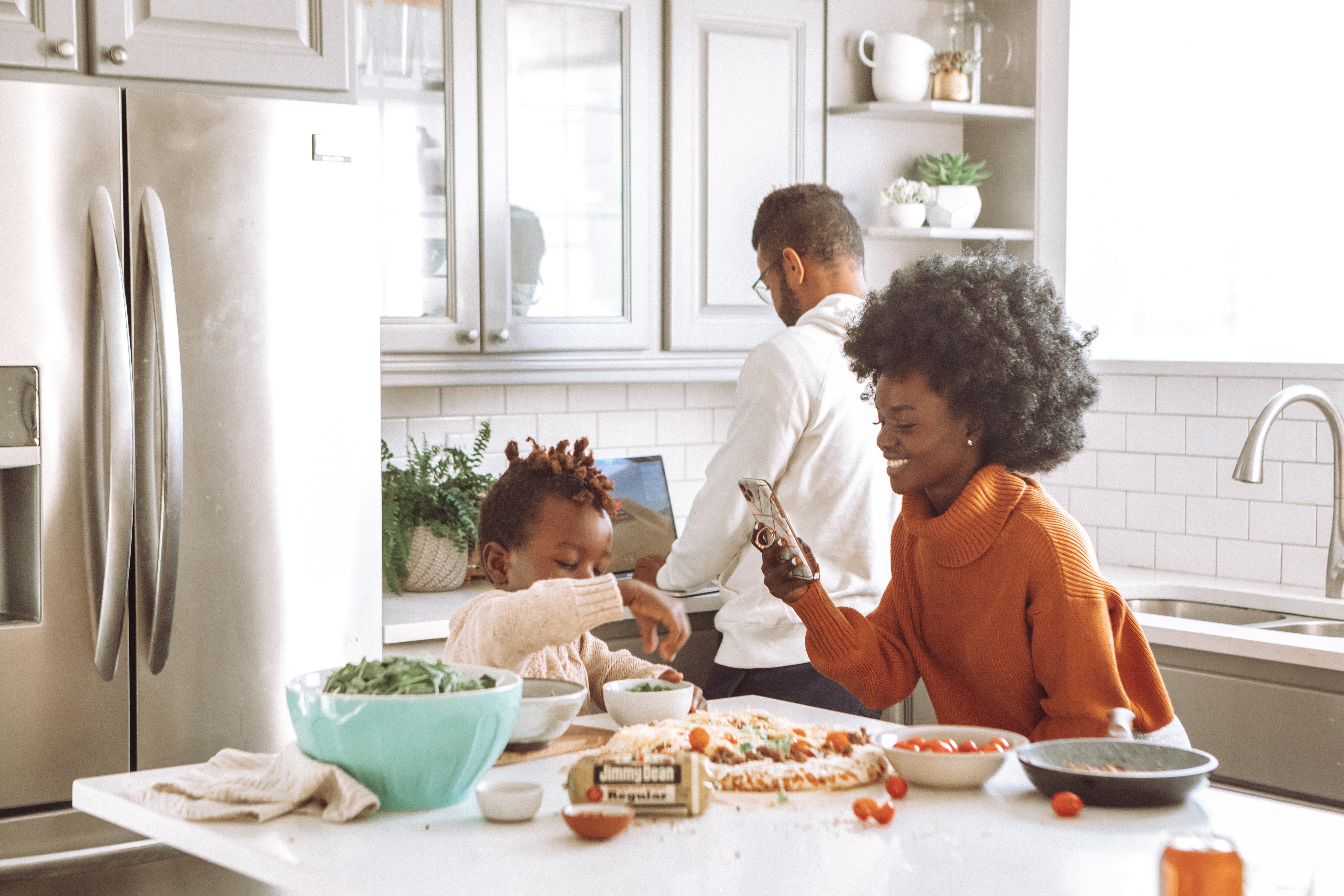 A mum taking a picture on her phone of her son making a pizza in the kitchen.