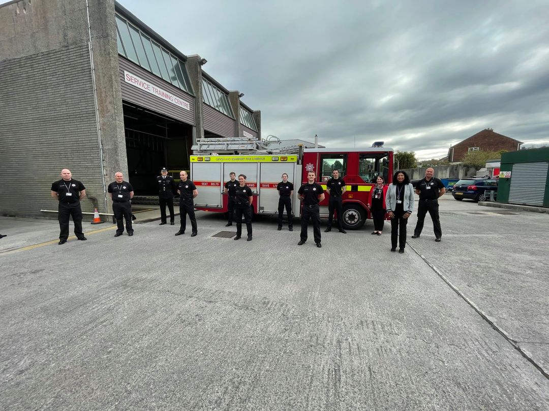 Twelve members of the Service stood in front of a fire appliance on a grey cloudy day. The six in the middle are the on-call apprentices, in their black uniform. Other firefighters are stood to the left and right, as well as two female support staff.