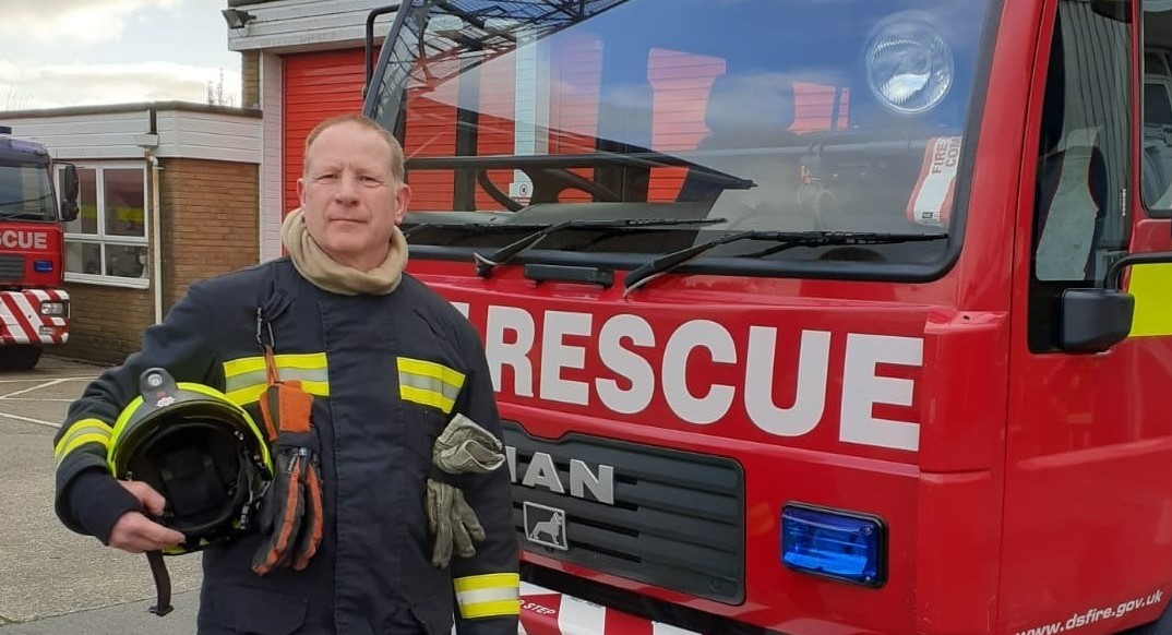 Community Officer Steve in his uniform, holding his helmet stood in front of a fire engine. He has short, buzzcut hair.