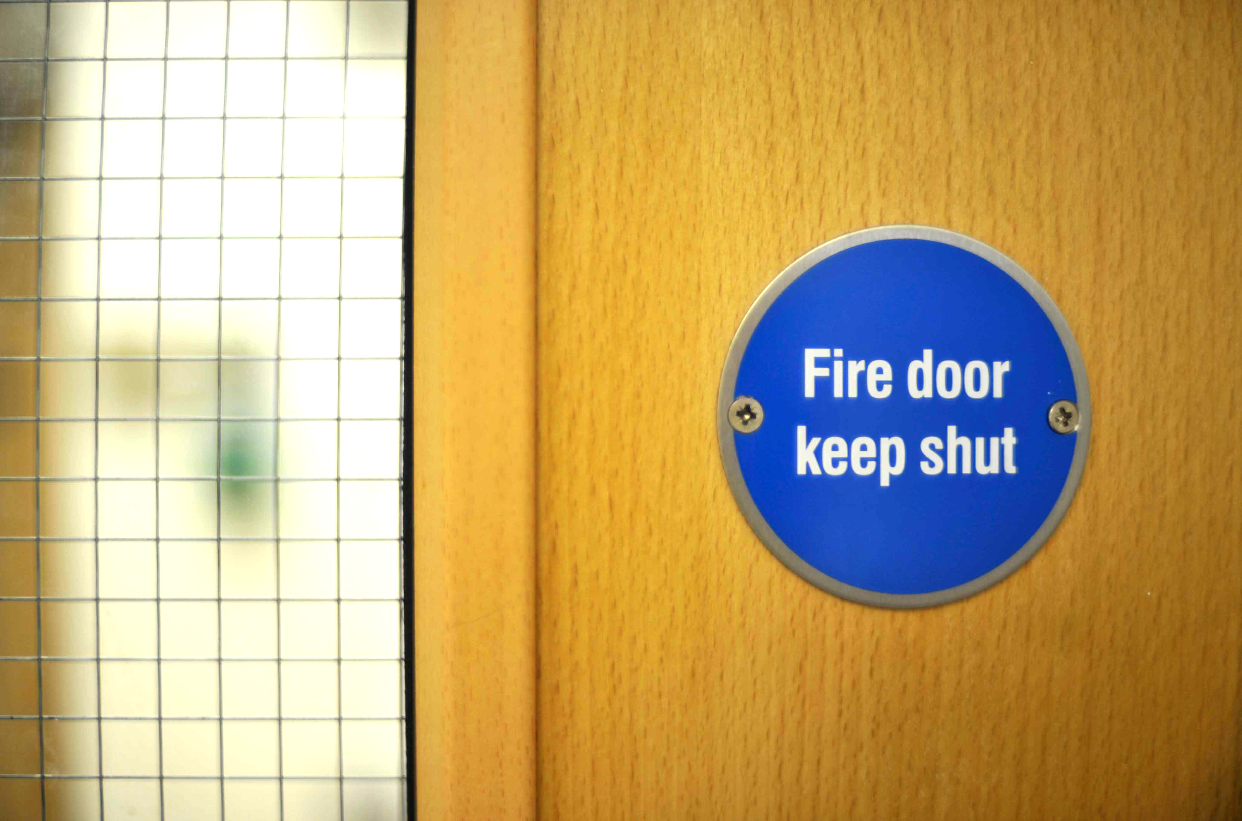 The blue circular sticker in a fire door which reads 'fire door keep shut' on a wooden fire door, with a but of the grid window of the fire door in the photo.