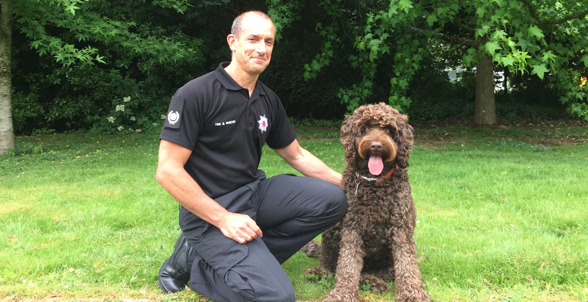 Digby the brown australian labradoodle sits on the grass next to his handler Matt Goodman. Matt is wearing the black Service uniform. 