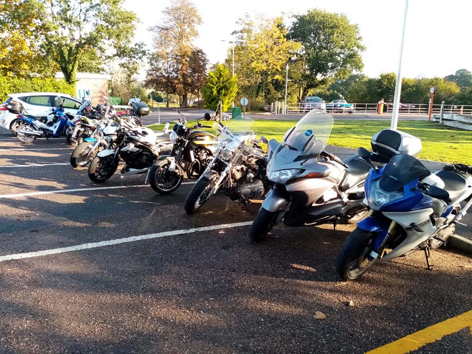 What looks to be about eight motorcycles parked in a line. They're various models and colours, from blue to silver to black. The background is carparks, where a few cars occupy some spaces, and grass and tall trees. It's a sunny day.