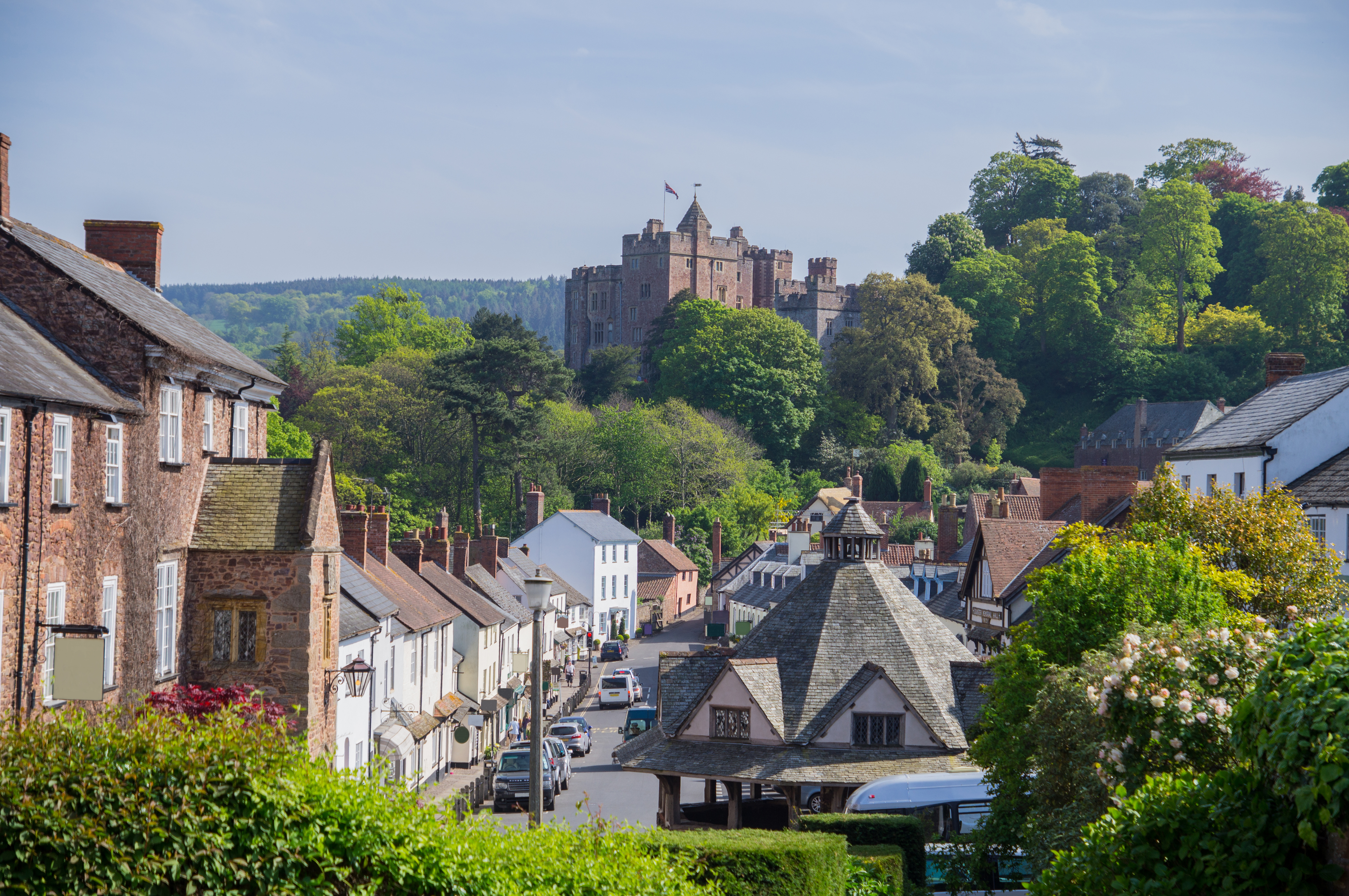 A street scene of Dunster village. It's a sunny day and a row of houses sits in front of Dunster Castle in the distance.