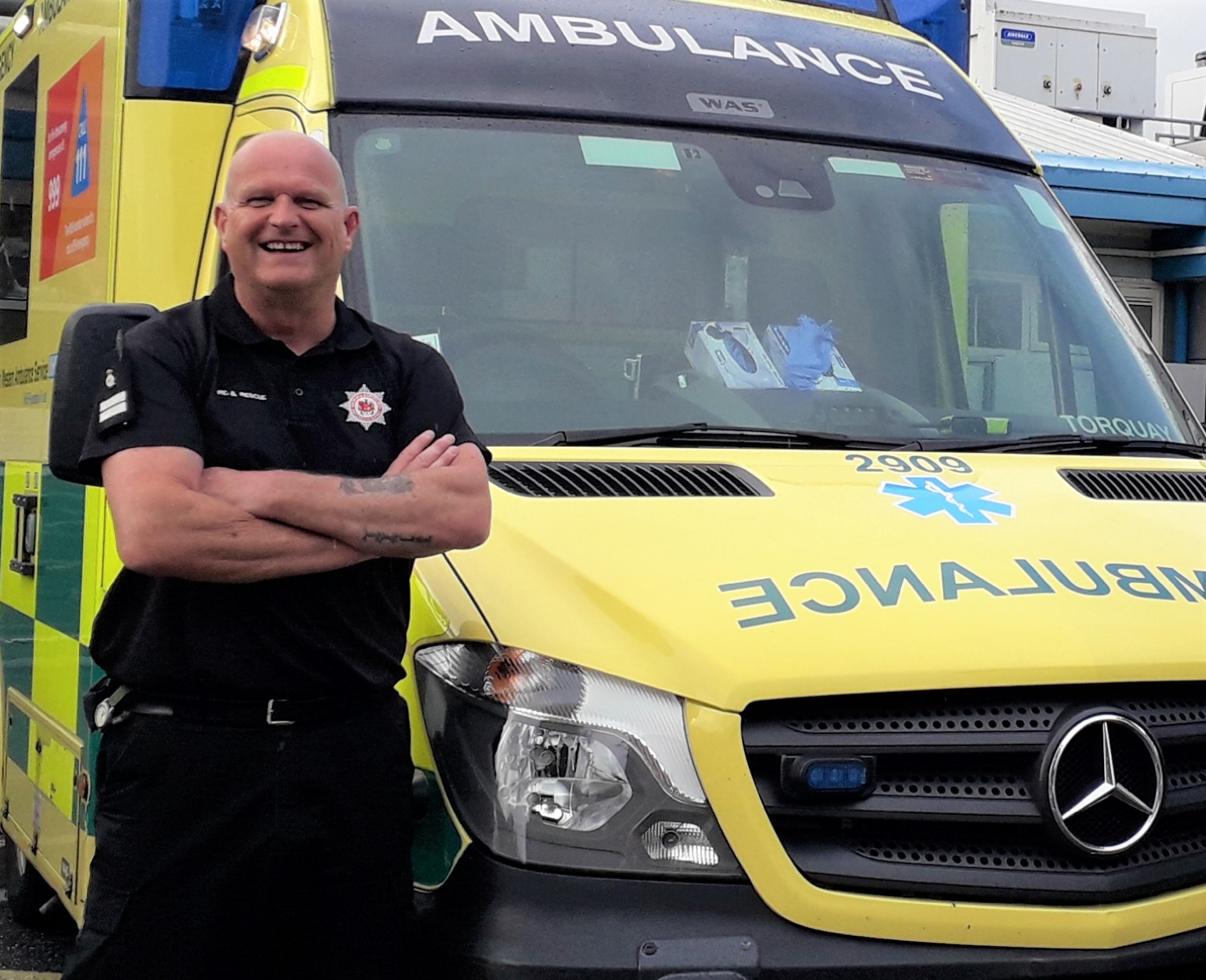 Our firefighter Kevin stood in front of an ambulance with his arms folded, smiling. He is in his fire service uniform (a black top with the logo and black trousers).