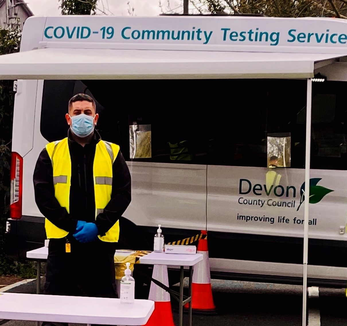 Our firefighter James stood in a mask, with gloves and a hi-vis jacket on in front of a Devon County Council testing van.