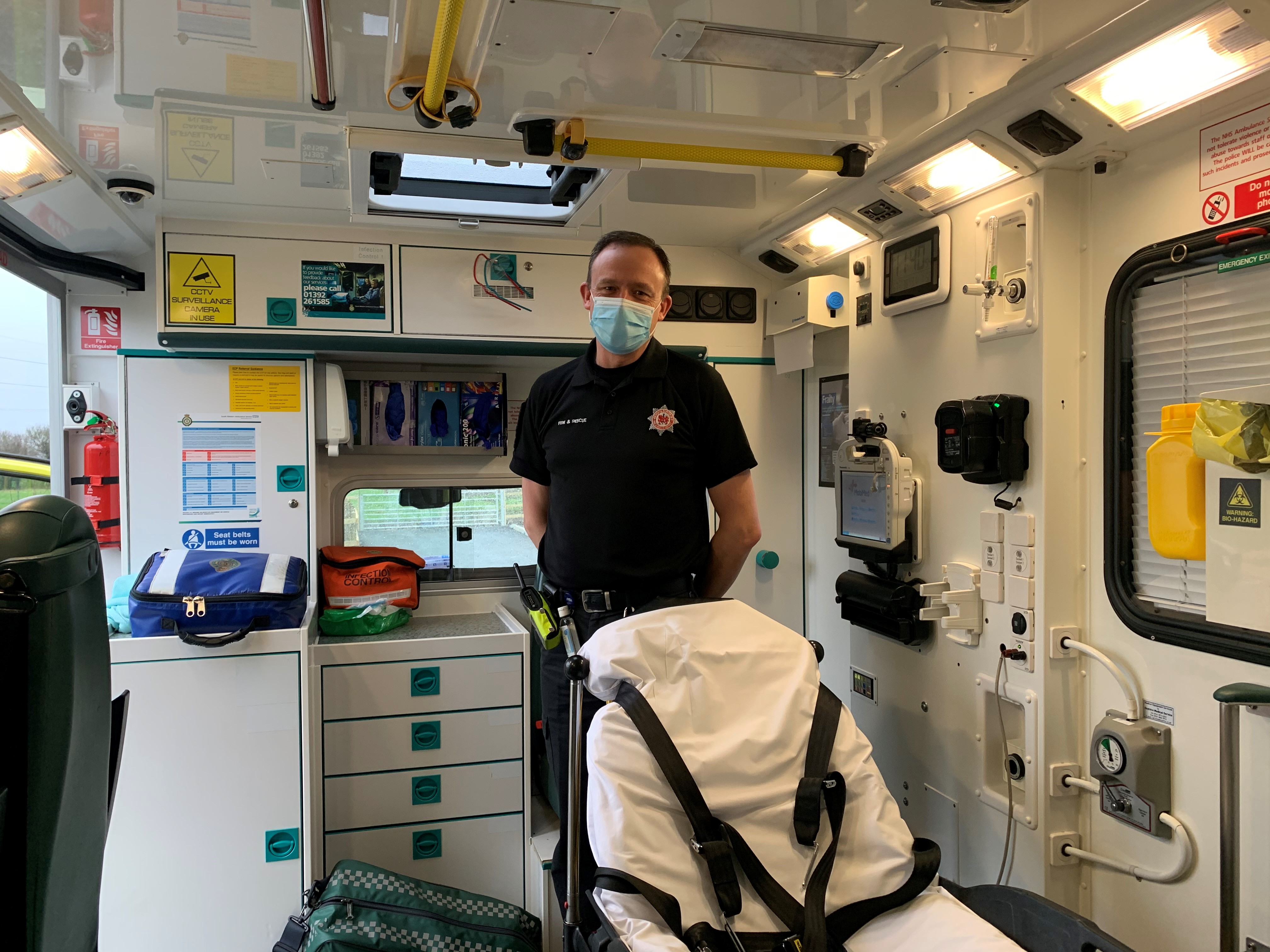 One of our firefighters, Robert Partridge, stood in the back of an ambulance surrounded by lots of ambulance equipment. He has his firefighter uniform on (which is a black shirt and trousers) and a mask.