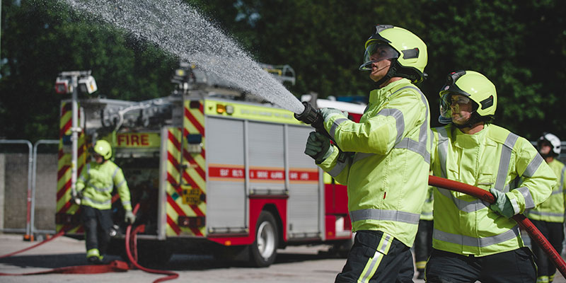 Two firefighters spray a jet of water from a hose. A fire engine is in the background.