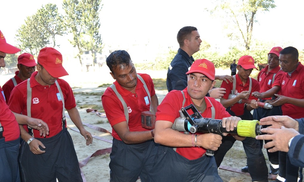 Firefighters being trained in Nepal. They're wearing their firefighter kit, which is all red, and a couple of them are holding hoses whilst the others nearby observe. One of our firefighters is stood amongst them, and one is just off camera helping.