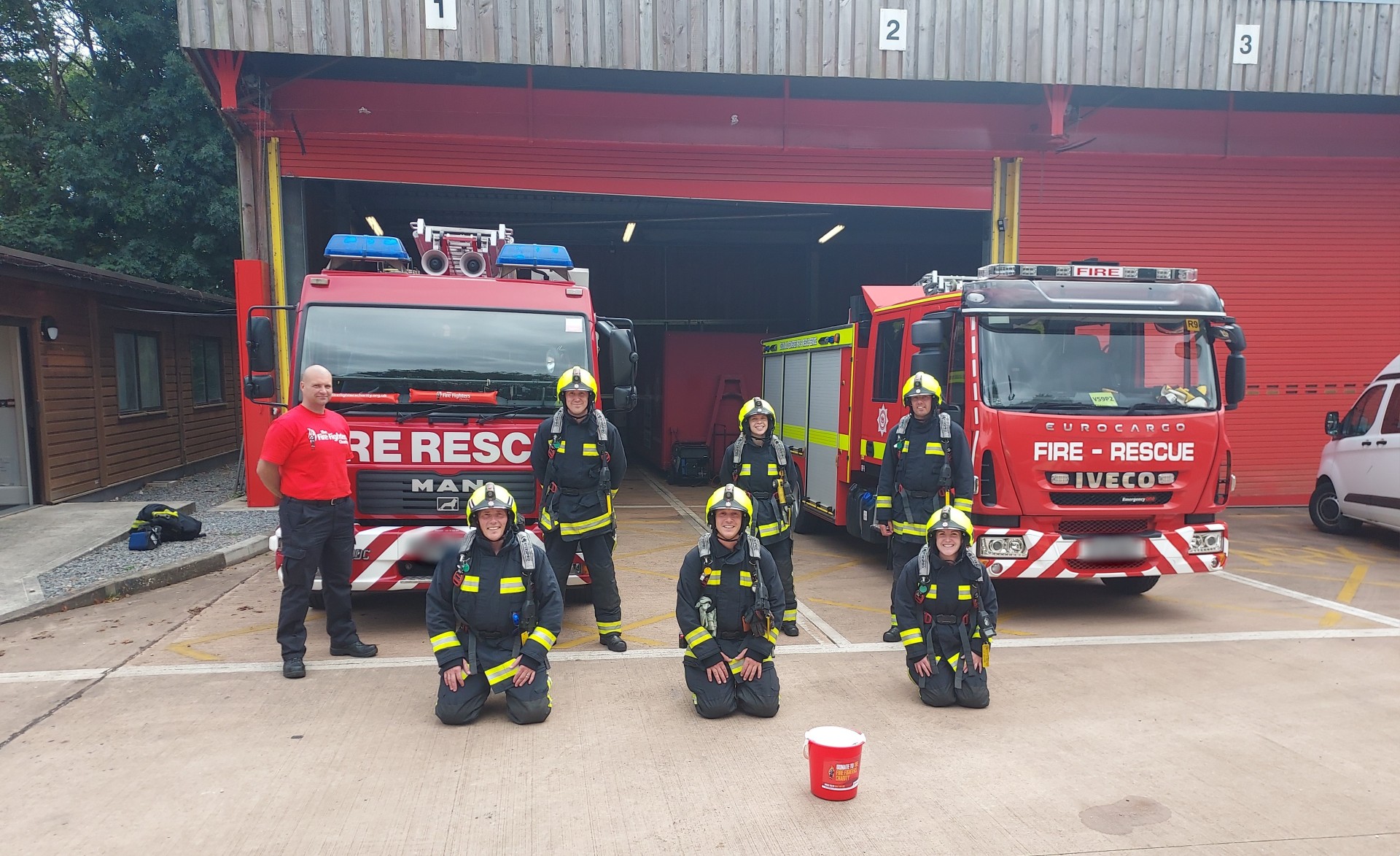 Seven people stood in front of two fire engines with an open station red garage behind them. Three are knelt on the ground, and four are stood behind. One is in a red Fire Fighters charity shirt whilst the others are in full kit. 