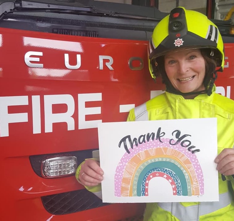 Sally stood in front of a fire engine in her firefighter kit, which is a neon jacket and matching helmet. She's smiling and holding an A4 piece of paper which has 'thank you' written on it alongside a rainbow with purple, orange, green, blue and red.