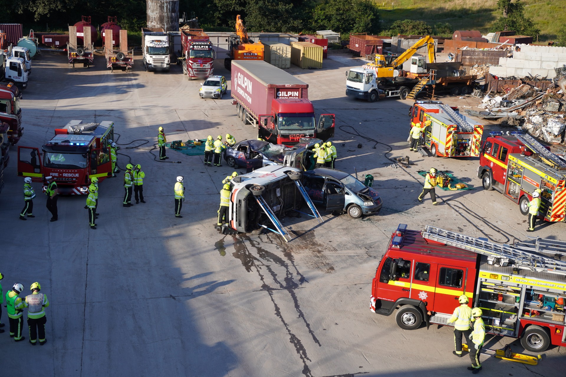 A concrete space being used for road safety training. There are lorries around the edge, four fire engines, firefighters dotted around and a model of a road traffic collision in the middle between three cars, a van and a lorry. A police car is also nearby.