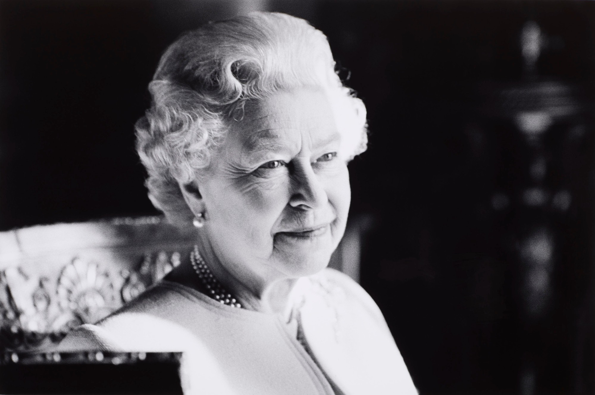 Her Majesty The Queen is photographed sitting down and looking off to the side of the camera in a black and white photograph.