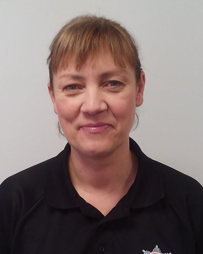 A woman with light brown hair stands in front of a grey background. She is smiling and wearing a black Service polo shirt.