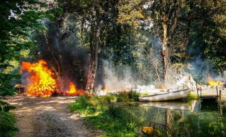 A bright, burning caravan fire spreading through a green forest by a small pond.