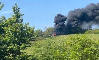 A photo of an out of control bonfire spreading across a hedgerow in field, with lots of black smoke floating up into the sky.