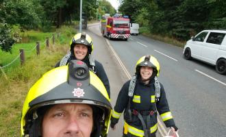 Three firefighters walking on the pavement together in full kit, smiling for a selfie. The road can be seen next to them, with a fire engine driving nearby as well as two white vans on the other side of the road.