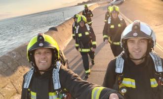 Seven firefighters in full kit walking along Exmouth beach just after sunrise. The sea can be seen next to them. They are walking in pairs, one behind the other.