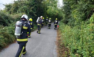 Seven firefighters walking along a lane lined by bushes and trees. It's a photo taken from behind, so their breathing apparatus is visible.