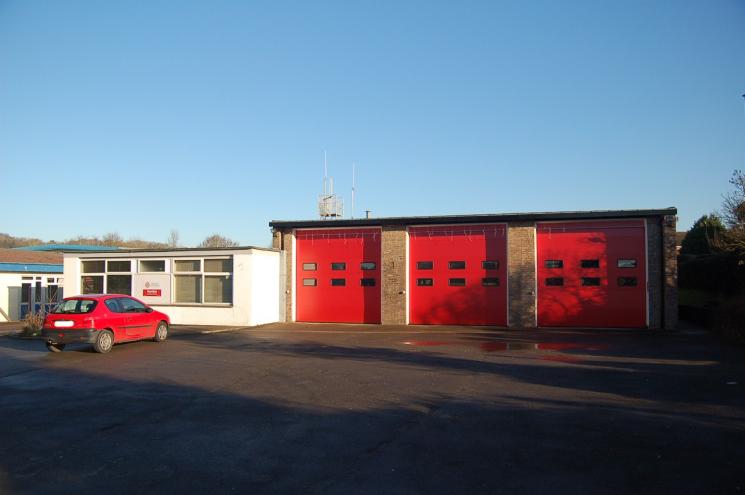 A photo of Honiton Fire Station's three red garages and small white building, with a red car parked outside.
