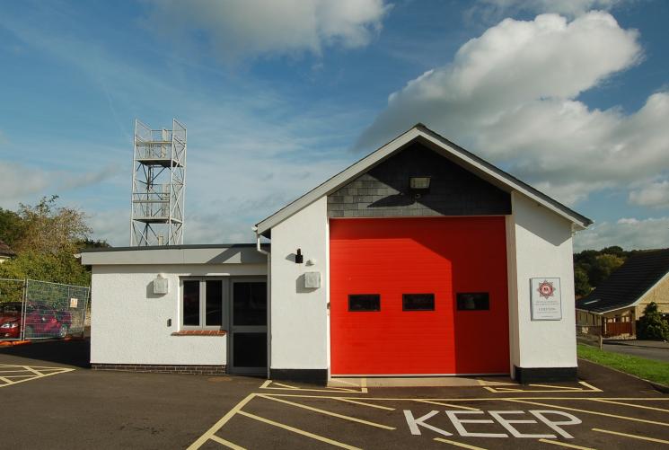 Colyton Fire Station's white building on a sunny day, with a small entrance door on the left and one garage with a red door on the right, where a fire engine is inside.