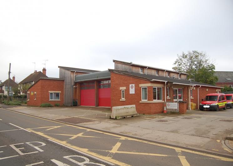 A photo of Crediton Fire Station - two red garages inbetween wood and brick buildings, and two red vans parked at the side.