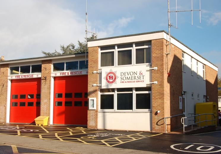 A photo of Sidmouth Fire Station, which has two red garages attached to a brick and white building, with the Service logo in the middle.