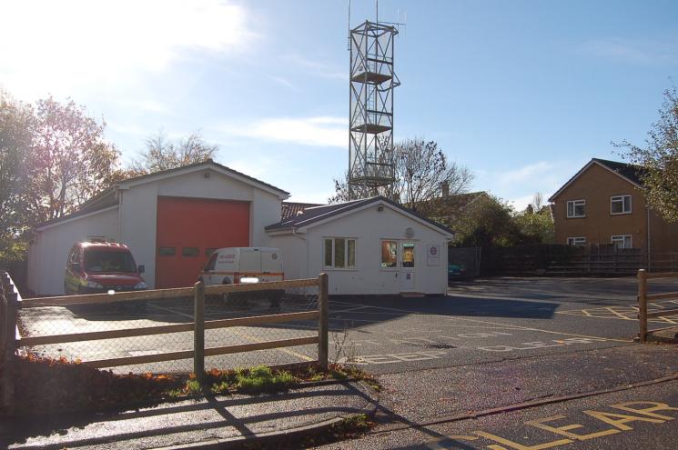 A photo of Seaton Fire Station, with a white entry building and one red garage, and two vans - one red and one white - parked outside.