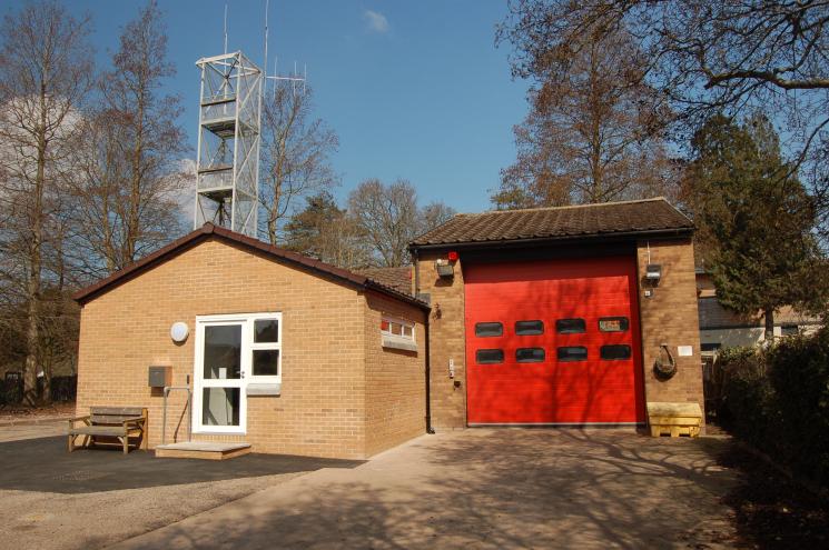 A photo of Ottery St Mary Fire Station's small entrance and one red garage, surrounded by trees on a sunny day.