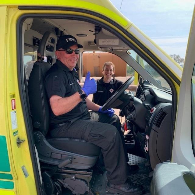 A photo of two of our ambulance driving volunteers, one behind the ambulance's wheel with gloves on and a thumbs up, the other behind him stood outside of the vehicle smiling.