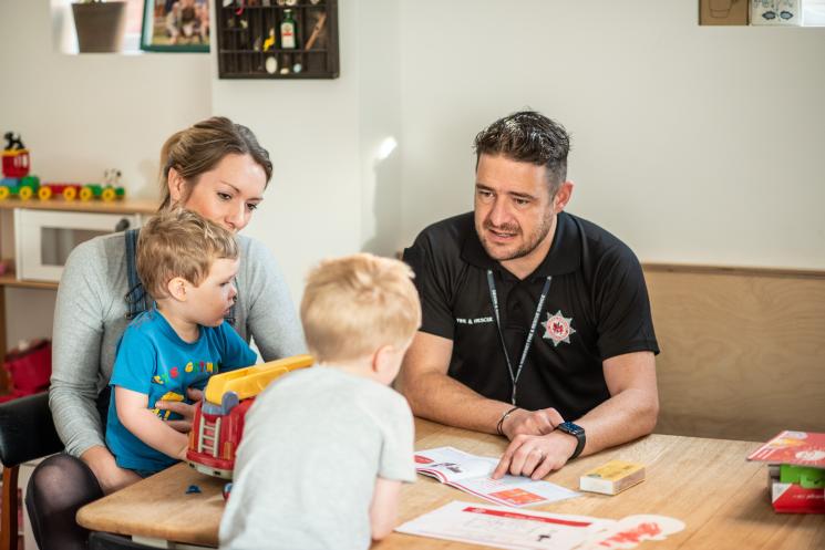 A mother sits with her two children and a home safety technician at their kitchen table. They are talking about fire safety with a toy fire engine on the table. 