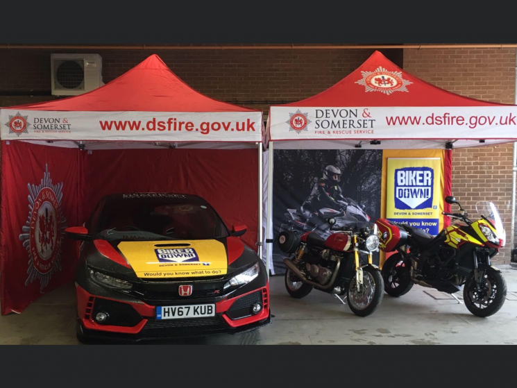 A honda car and two motorcycles are parked underneath a Devon and Somerset Fire and Rescue Service stand.