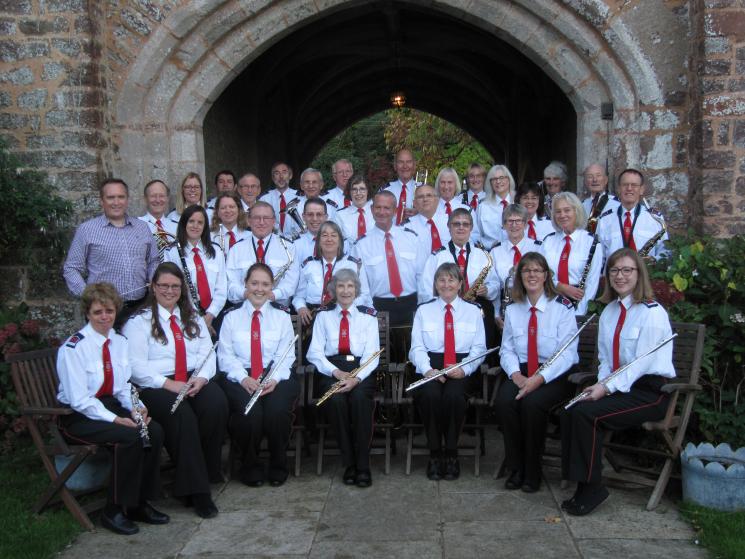 The Concert Band dressed in white shirts, red ties and black trousers holding their instruments. The front row are sat down while the other rows are stood.