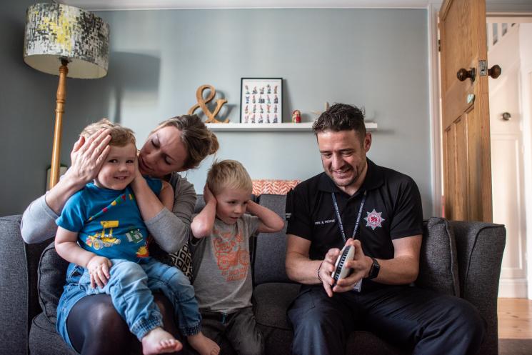 Home safety technician testing a smoke alarm by pressing the button. A mum and her two children are set next to him - the older child is covering his ears and the mum is covering the ears of the younger boy.
