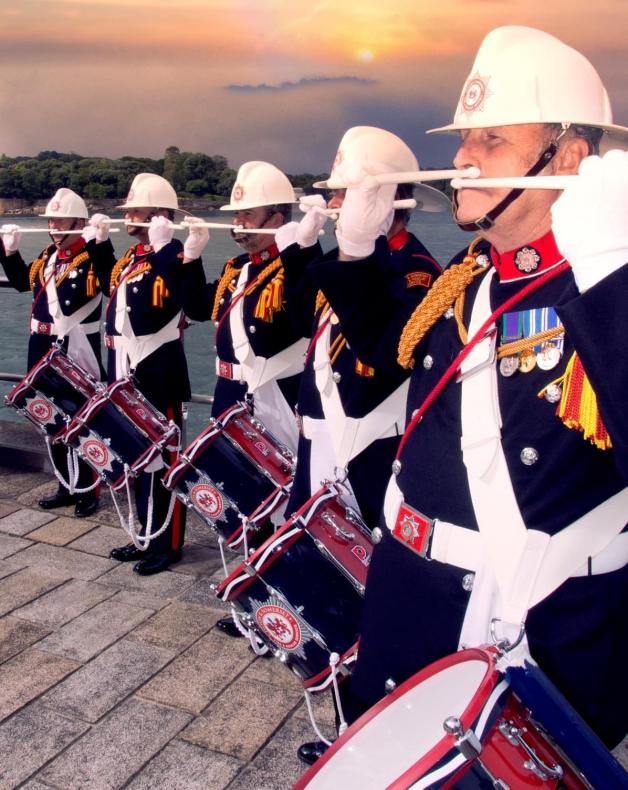 Five members of the Corps of Drums in full uniform, with white hats on. They have drums strapped to them and are holding the drumsticks under their noses. The background is beautiful sunrise over water.