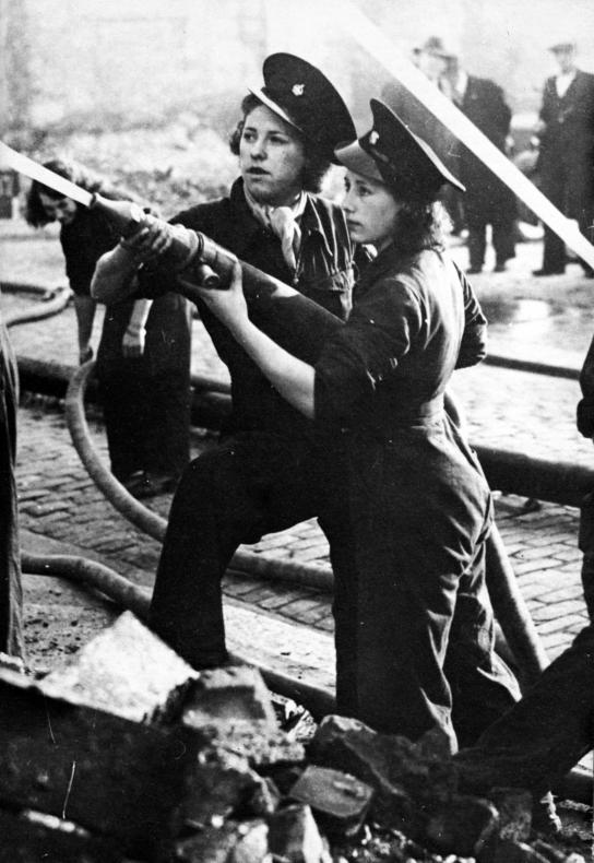 Two firefighting women holding a hose together in the street, in full uniform (jumpsuits and hats). They are aiming the water above them. Rubble can be seen at their feet.