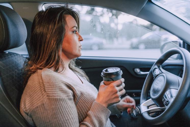 Woman with brown hair drinking a hot drink at the steering wheel.