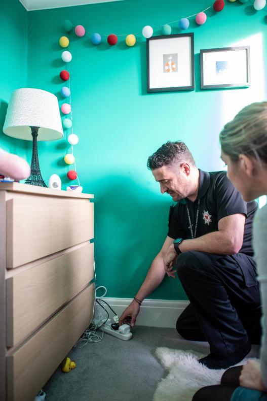 A home safety technician and woman knelt on the floor inside the woman's home, looking at extension leads and sockets on the carpet. There are lots of wires. The room is painted turquoise and there are round fairy lights on the wall. 