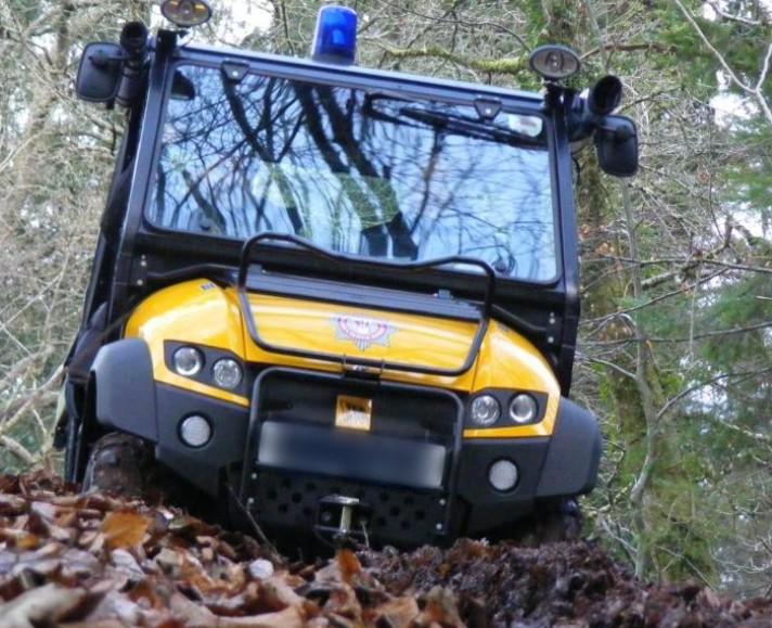 This is a small vehicle, with a blue siren on top and yellow bonnet and the Service logo. The rest is black, with a wide windscreen. It's seen in the photo travelling over muddy leaves in a woodland area with trees behind.
