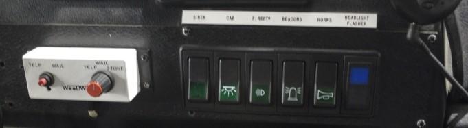 A white box with two red and black buttons that twist for different actions. This is next to a black panel of five rocker switches with green symbols. The switches are for acts such as sirens, horns and beacons. There is a black button next to these with a blue light which is labelled headlight flasher.