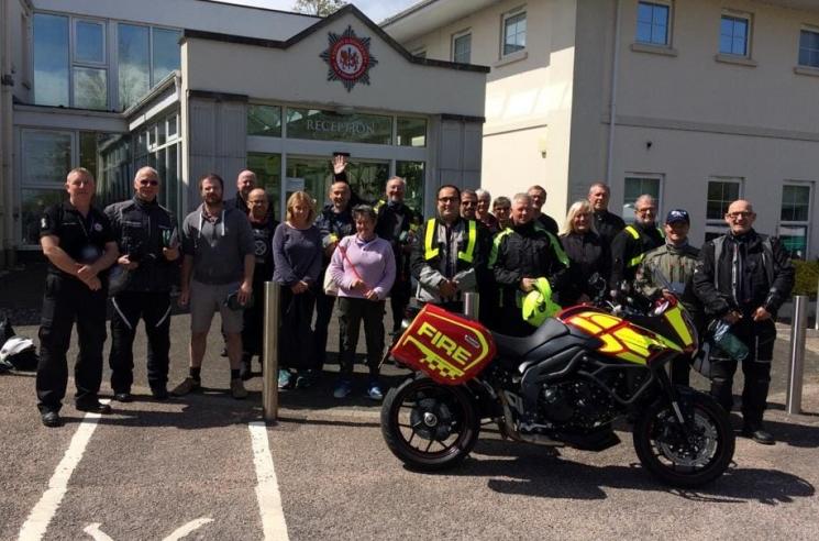 A group after a Biker Down event. There are 20 people stood together outside of Service Headquarters' entrance, which is glass with cream pillars and has 'Reception' written in white across the front. A red and neon yellow fire service motorcycle is in front of the group. It has fire written in capital neon yellow letters across the back.