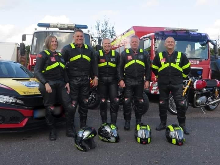 Five members of the Fire Bike team stood in full, matching motorcycle safety gear. Four helmets are all placed on the floor in front of them, and three Service vehicles - which are two engines and one car- as well as a motorcycle are behind them. The group from left to right is a woman, a man, another woman, and another two men. 