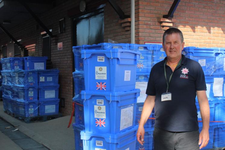 One of our firefighters, wearing a navy or black Service shirt and dark trousers, stood in front of lots of stacked, blue aqua boxes. The boxes are stacked in front of a brick building. 