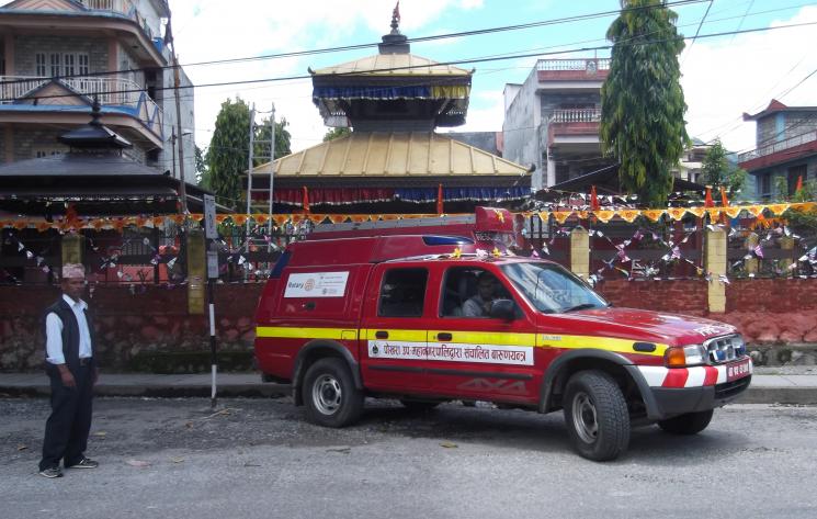 A firefighter from Nepal sat inside of a donated DSFRS Ford Ranger. The vehicle is red, with two doors each side to get into the seats and a storage area at the back. It has four wheels and a neon yellow line across the middle. Another man from Nepal is stood to the left of the vehicle. The vehicle is parked in a street, where there are flags and a decorated building behind.
