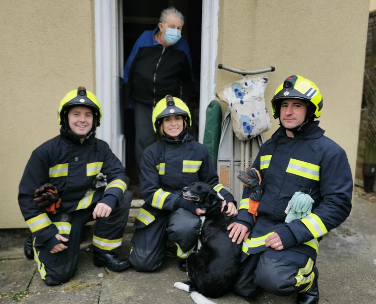 Three firefighters knelt down with Chi Chi the dog, who is black and white. Mr Green is stood in the doorway behind them.