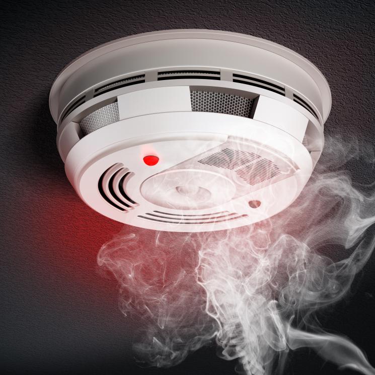 A smoke alarm on a dark ceiling, with a cloud of smoke rising towards it.